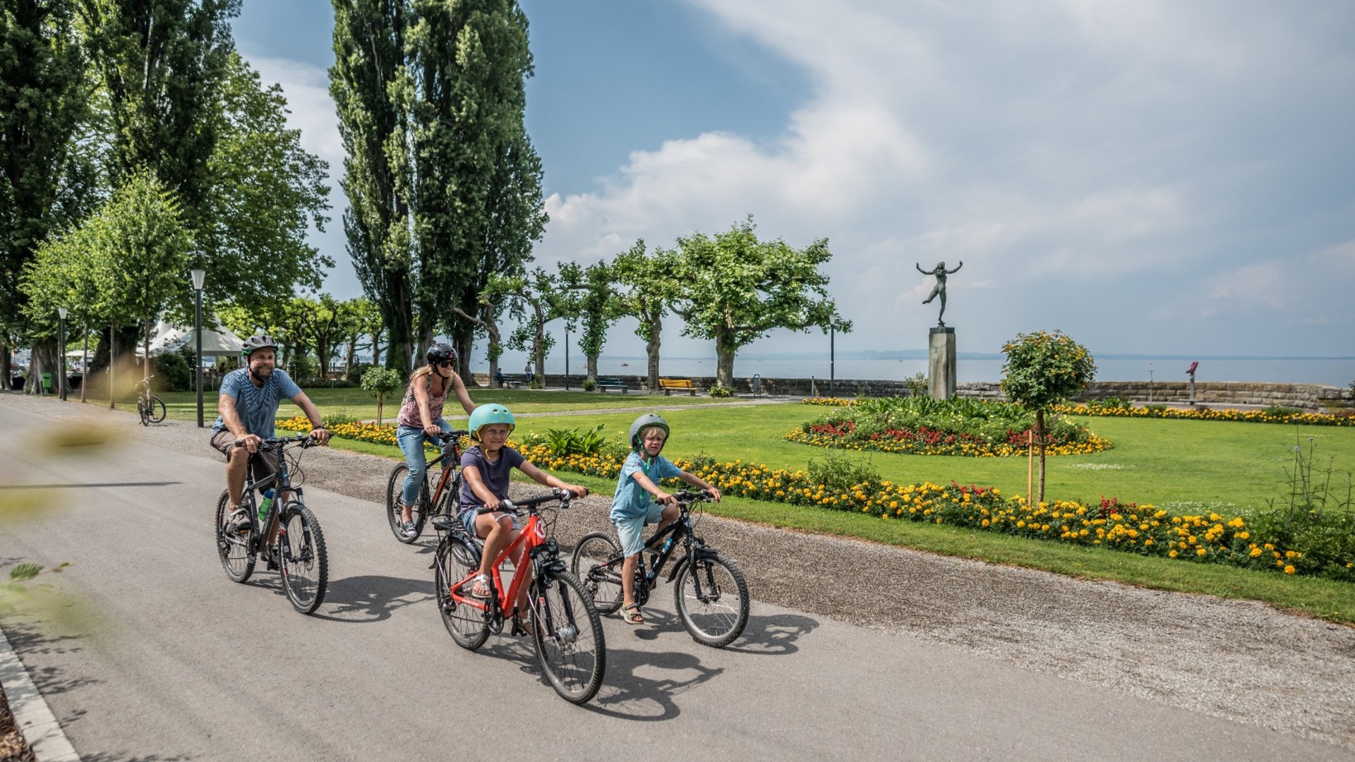 Family cycling on a path by a lakeside park with a statue