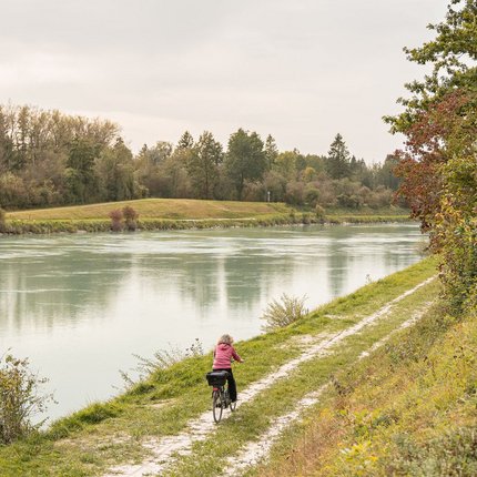 Person cycling along a narrow path by a river with trees