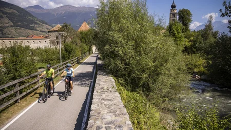 Ein Genuss-Radtag am Reschensee © Velontour - Heiko Mandl Zwei Radfahrer auf einem Weg neben Fluss und historischen Gebäuden in den Bergen