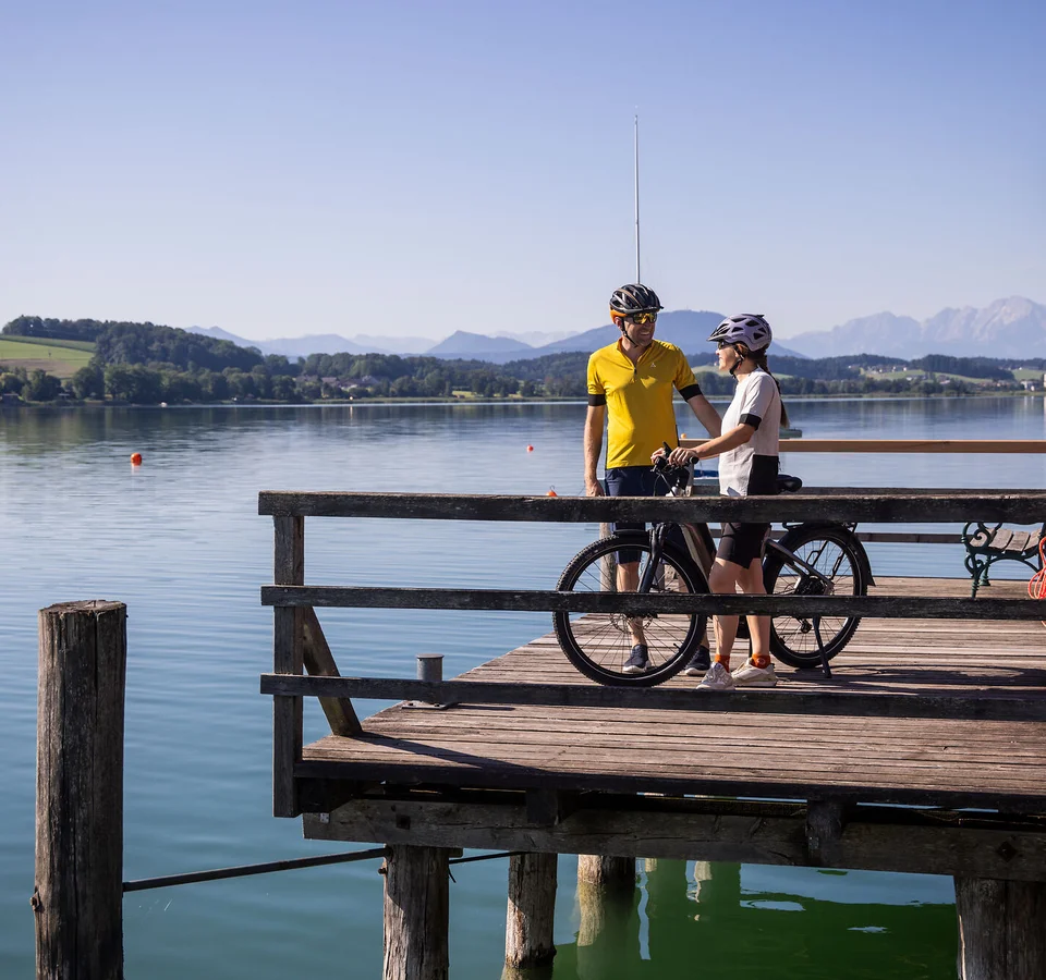 Zwei Radfahrer auf einem Steg am See mit Bergen im Hintergrund