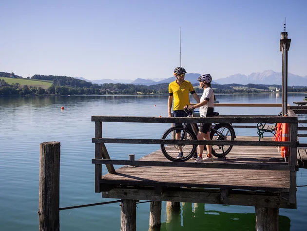 Zwei Radfahrer auf einem Steg am See mit Bergen im Hintergrund