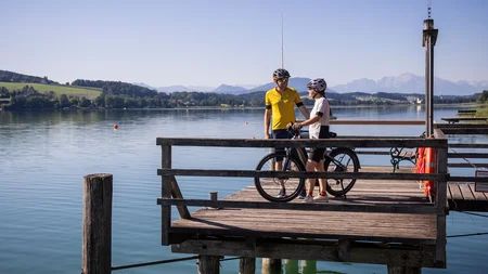 Zwei Radfahrer auf einem Steg am See mit Bergen im Hintergrund