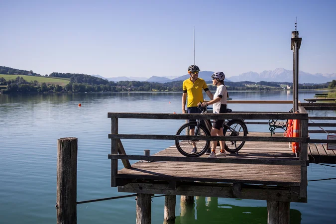 Zwei Radfahrer auf einem Steg am See mit Bergen im Hintergrund