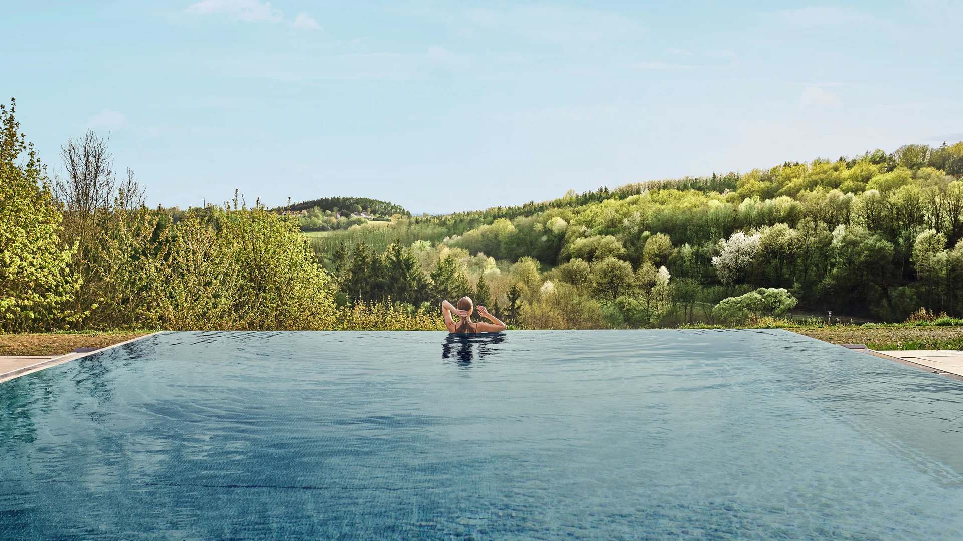 Person entspannt im Infinity-Pool mit Blick auf bewaldete Hügel