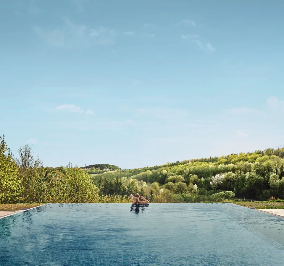Person entspannt im Infinity-Pool mit Blick auf bewaldete Hügel