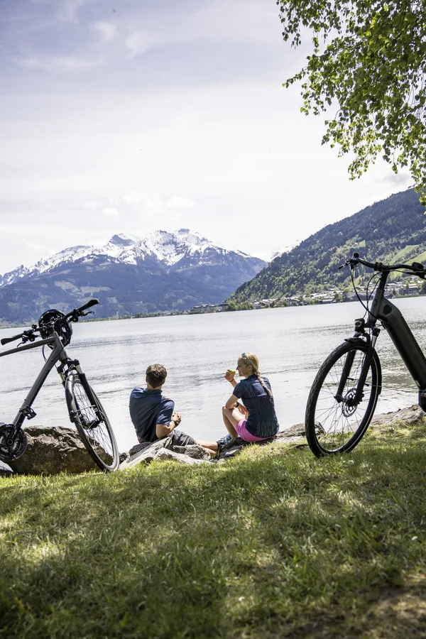 RadGENUSS im kleinen Juwel Maishofen © Velontour Pärchen sitzt am Seeufer mit Fahrrädern und Blick auf verschneite Berge