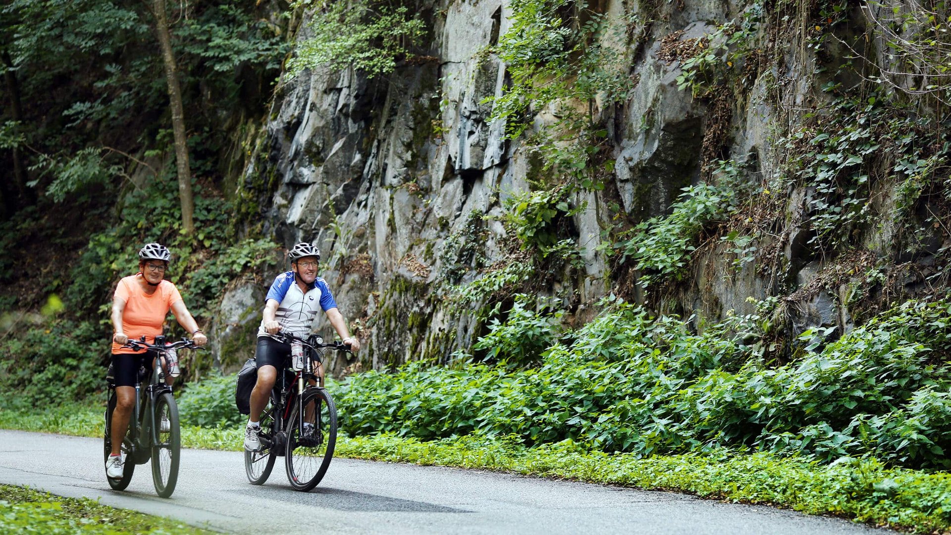 Two cyclists riding on a road beside a rocky wall with greenery
