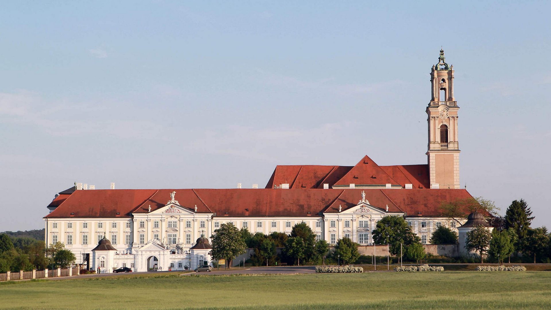 Monastery with red roof and tower in front of green field under blue sky
