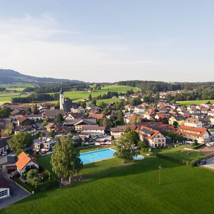 Haunsbergrunde © Jakob Eder Luftaufnahme eines Dorfes mit Schwimmbad und grünen Wiesen unter blauem Himmel