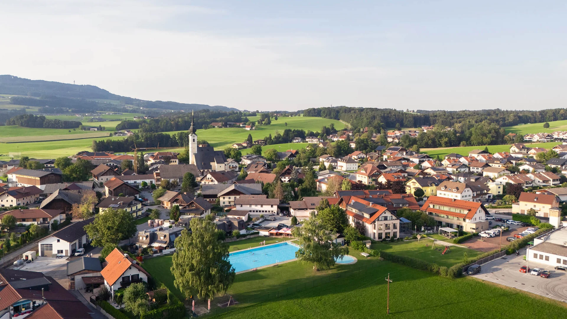 Haunsbergrunde © Jakob Eder Luftaufnahme eines Dorfes mit Schwimmbad und grünen Wiesen unter blauem Himmel