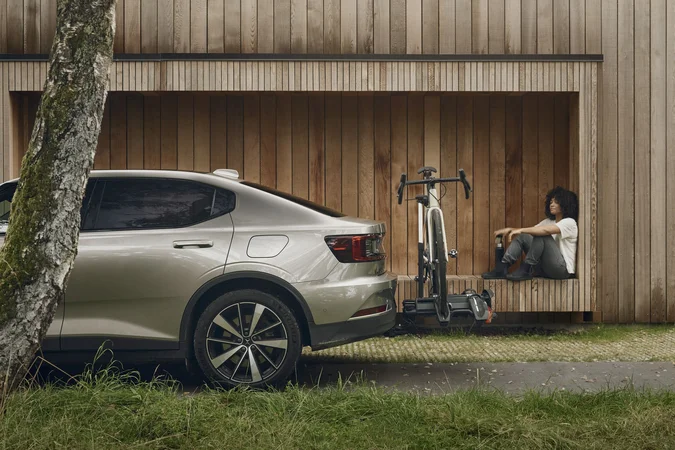 Person sitzt neben einem Fahrrad an einer Holzfassade, daneben steht ein Auto