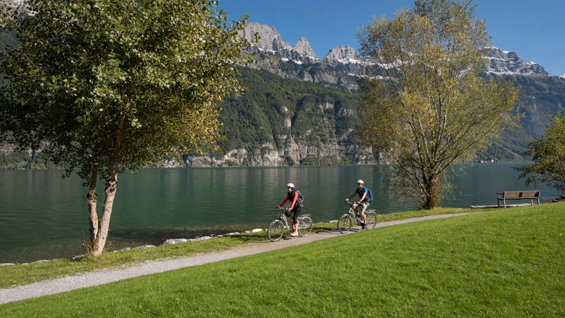 Zwei Radfahrer auf einem Weg am See mit Bergen und Bäumen