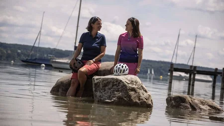 Zwei Frauen mit Fahrradhelmen sitzen auf Steinen im Wasser an einem See