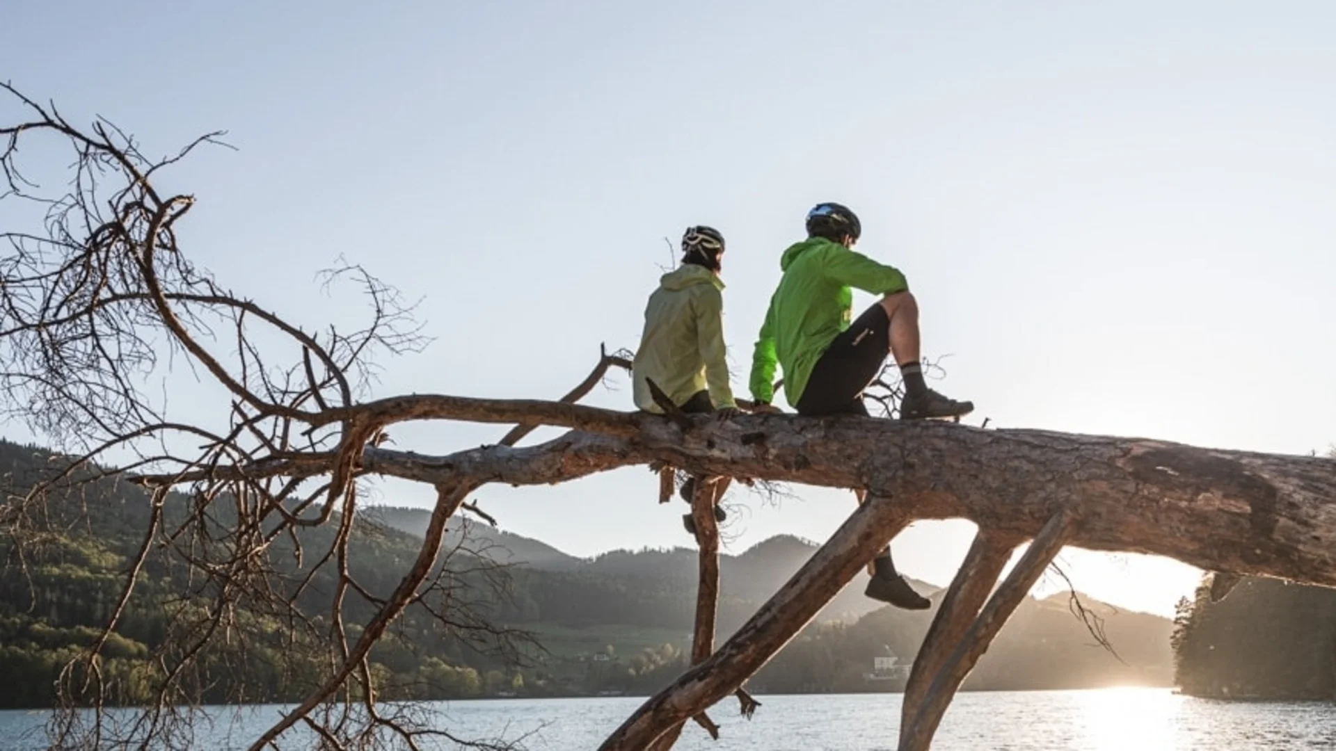 Two cyclists sitting on a tree above lake at sunset