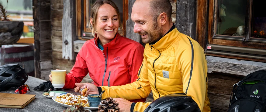 Paar in Outdoor-Bekleidung isst und trinkt an Holztisch vor Hütte