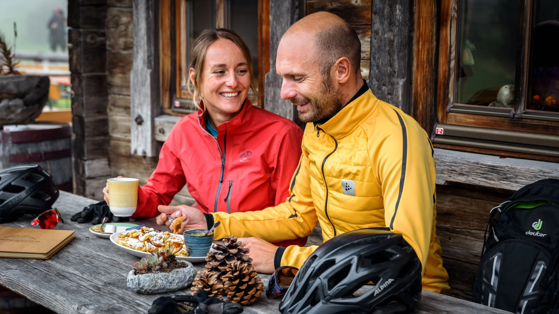 Paar in Outdoor-Bekleidung isst und trinkt an Holztisch vor Hütte