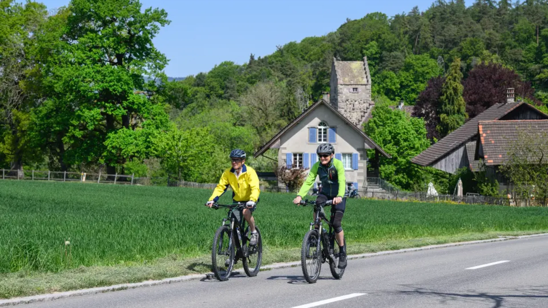 Tour-Detail © Balz Kubli Zwei Radfahrer fahren auf einer Landstraße mit grünen Wiesen und Häusern im Hintergrund
