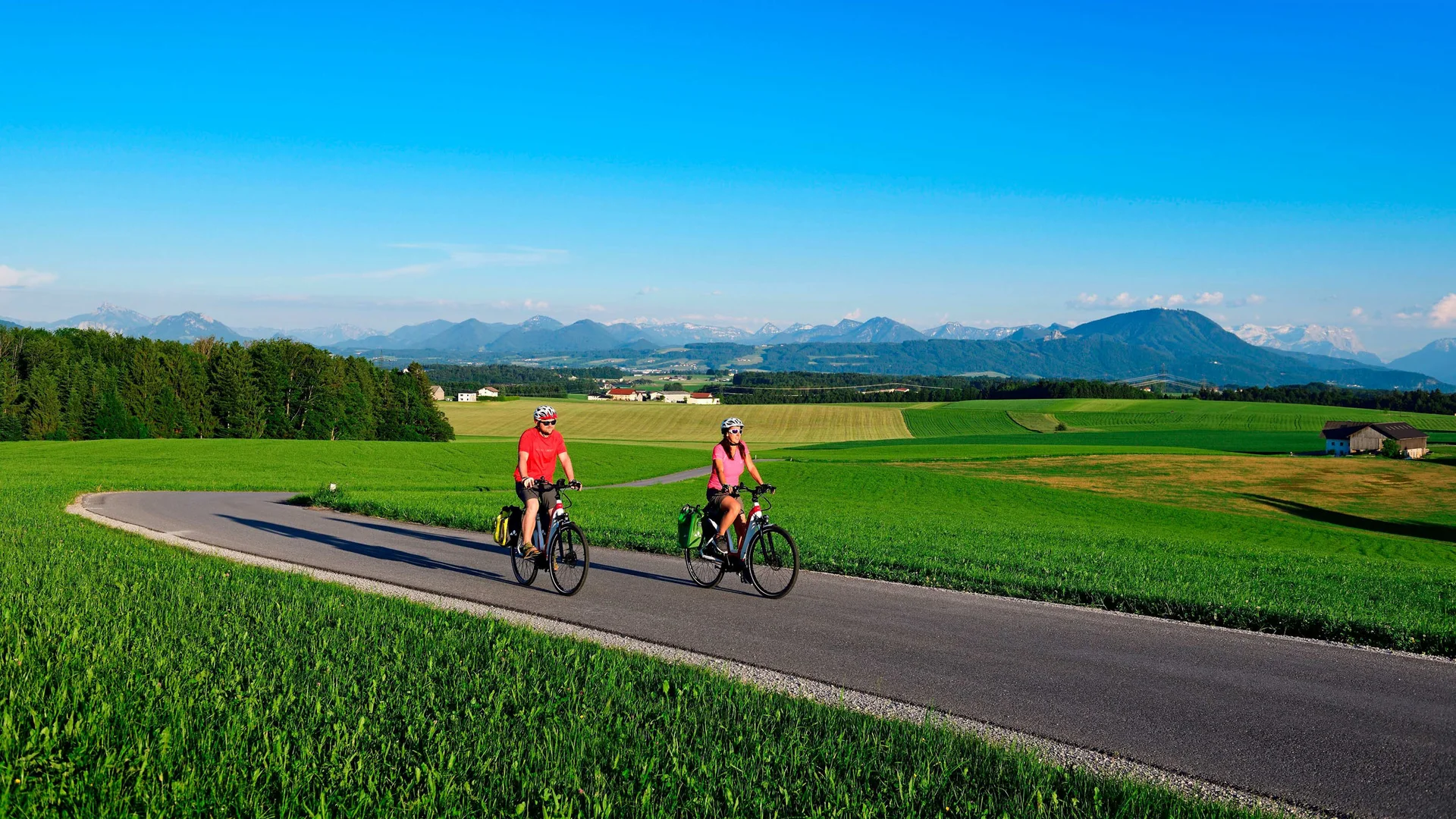 Two cyclists ride on a country road through green fields with mountains in background
