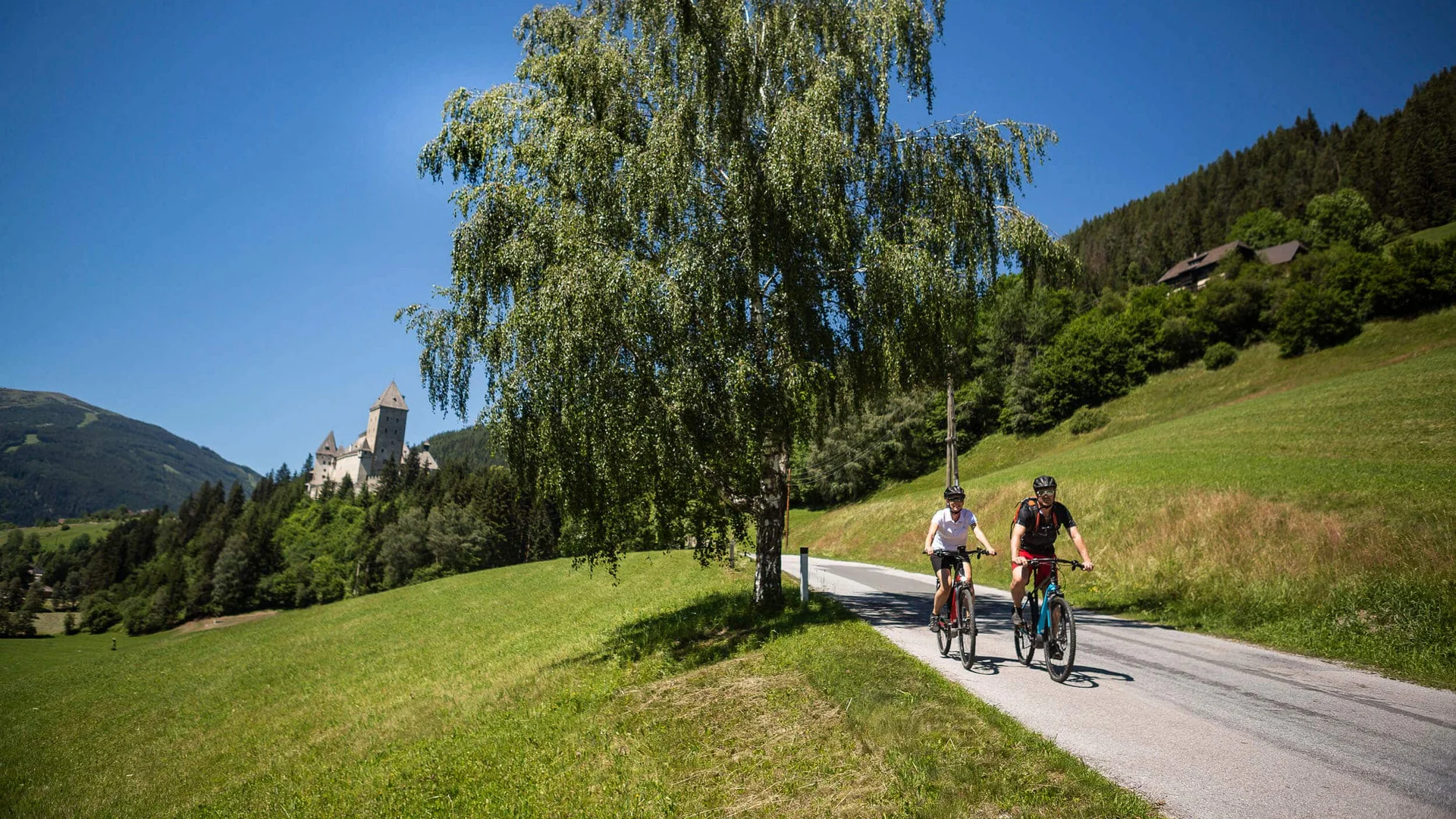 Two cyclists on country road in green landscape with castle in background