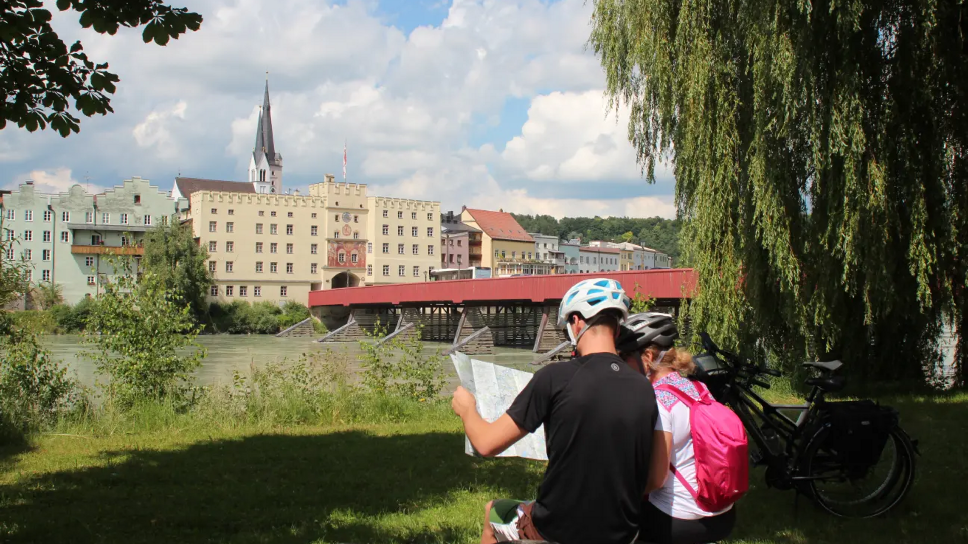 Two cyclists reading a map by river with red bridge and historic buildings