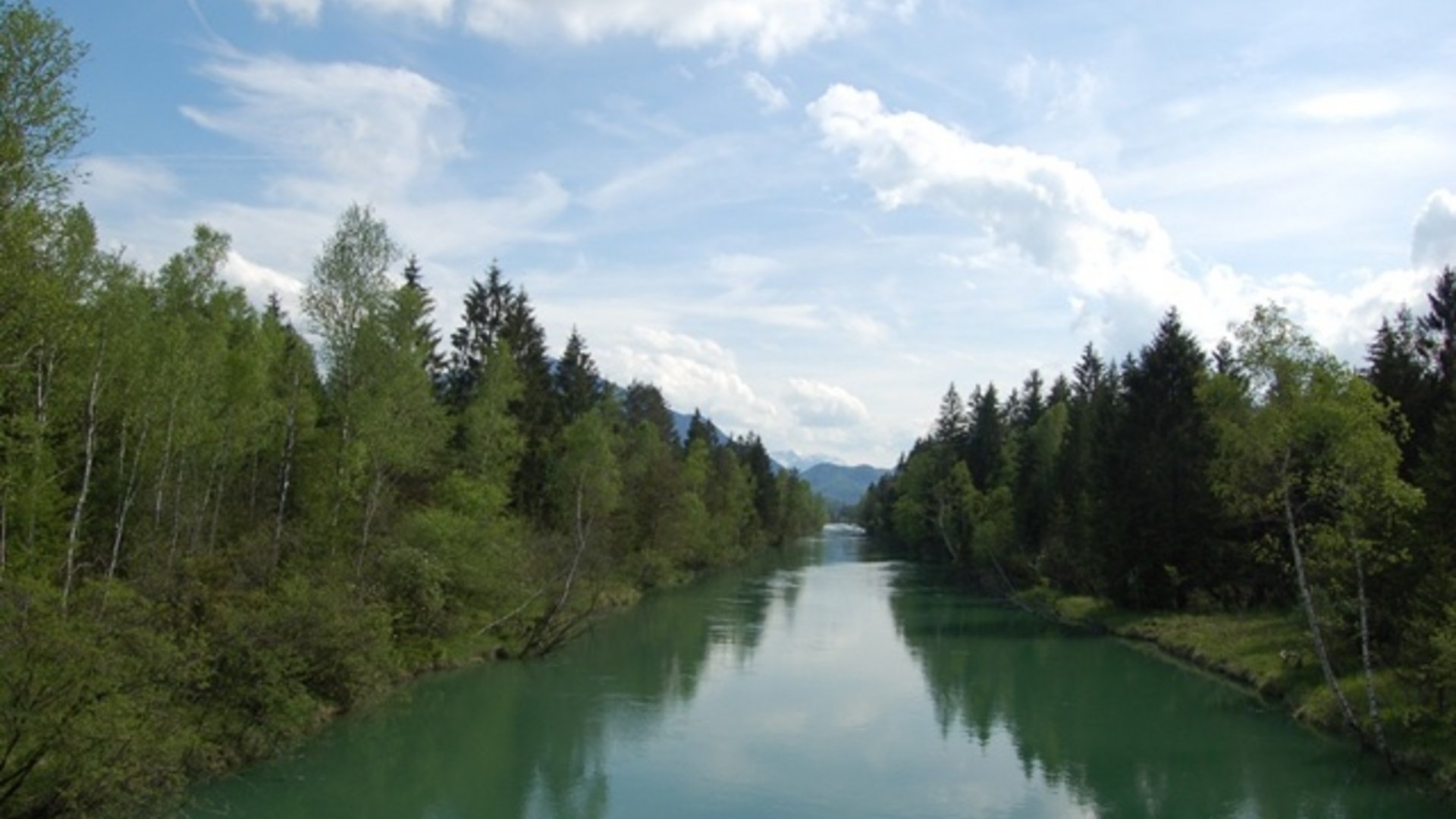 Green river surrounded by trees and mountains under a blue sky with clouds