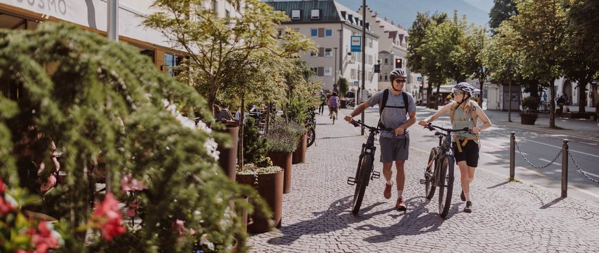 Two cyclists wearing helmets walking their bikes on a cobblestone street in a city