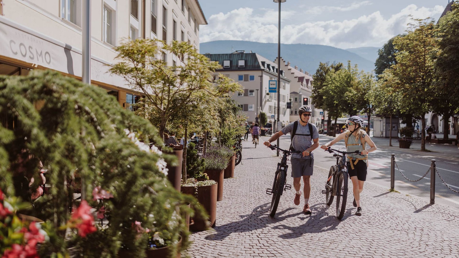 Two cyclists wearing helmets walking their bikes on a cobblestone street in a city