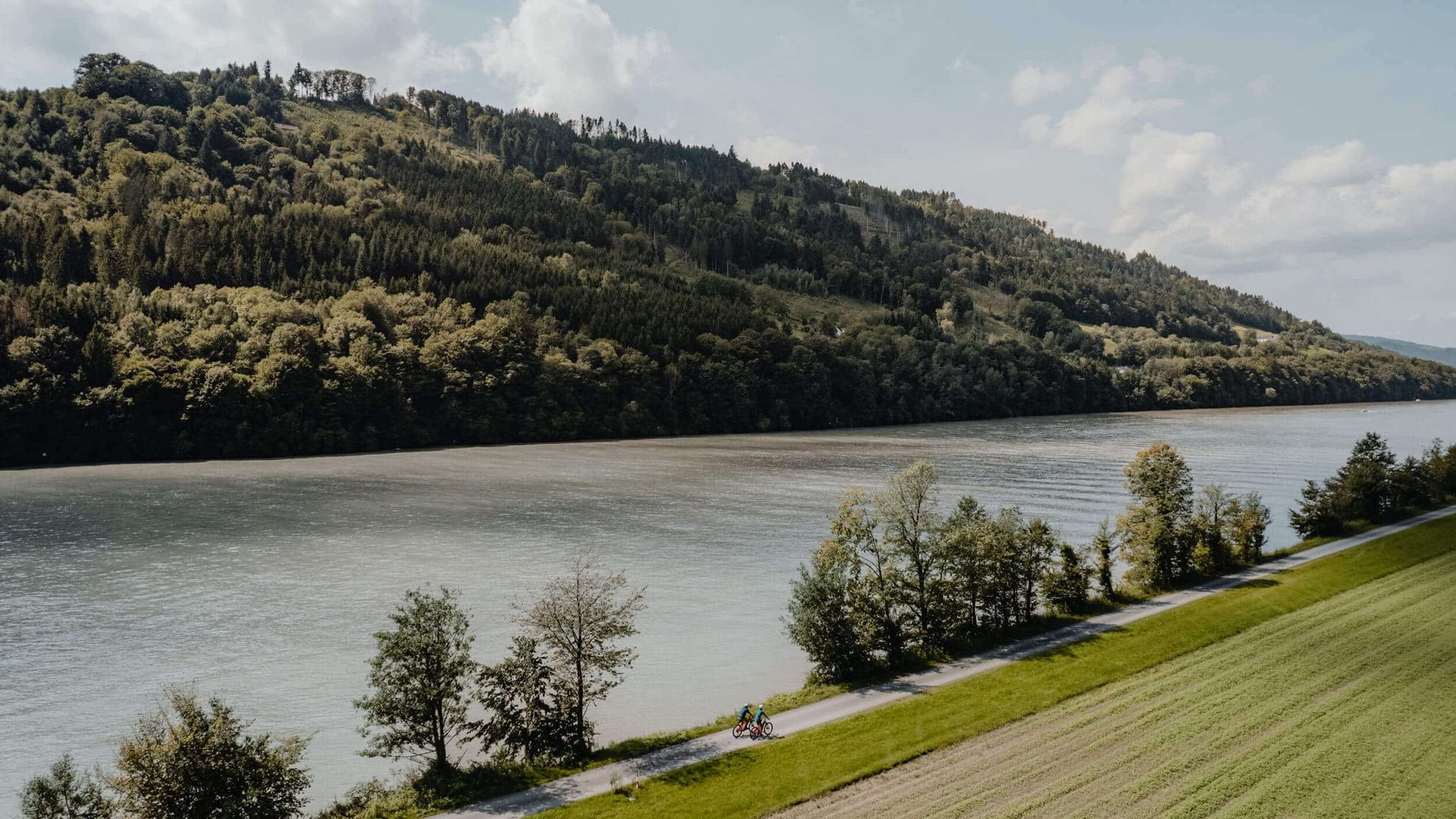 Cyclist on a path alongside a river with forested hills in the background