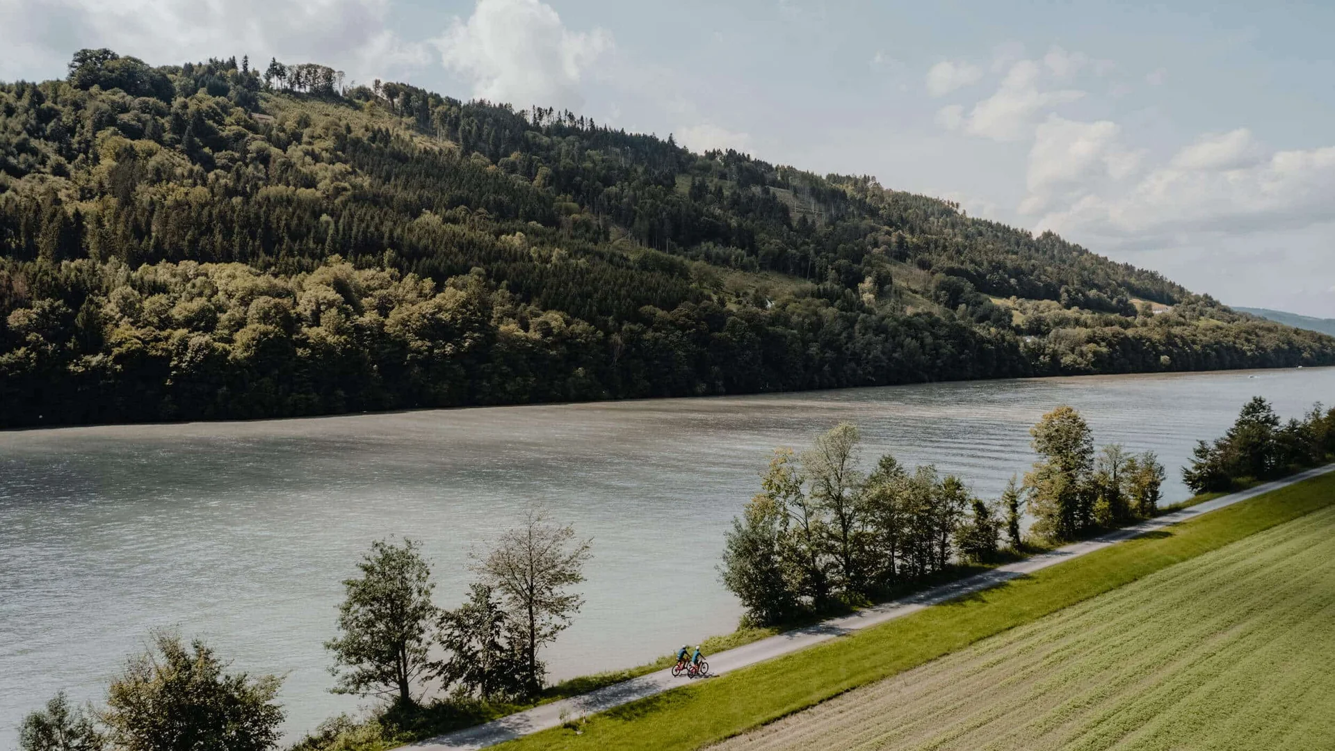 Fahrradfahrer auf einem Radweg neben einem Fluss und bewaldeten Hügeln