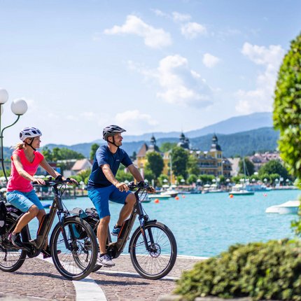 Paar fährt mit E-Bikes an einem See in einer sommerlichen Stadt
