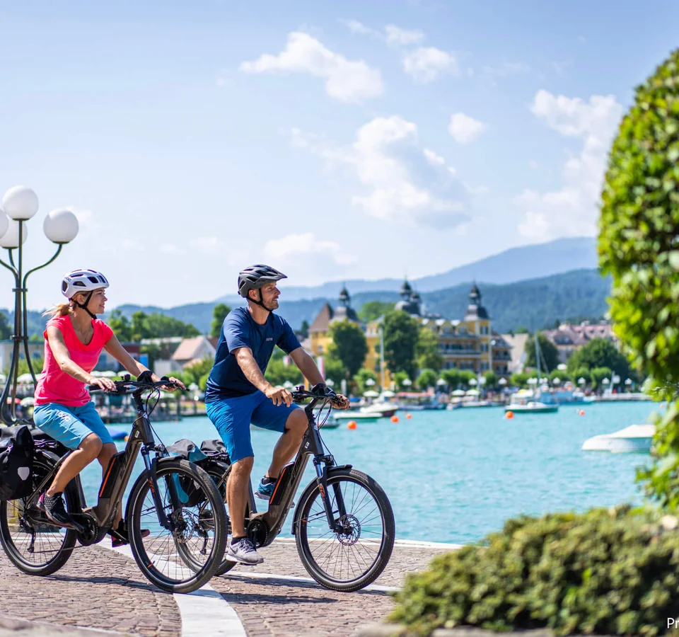 Paar fährt mit E-Bikes an einem See in einer sommerlichen Stadt