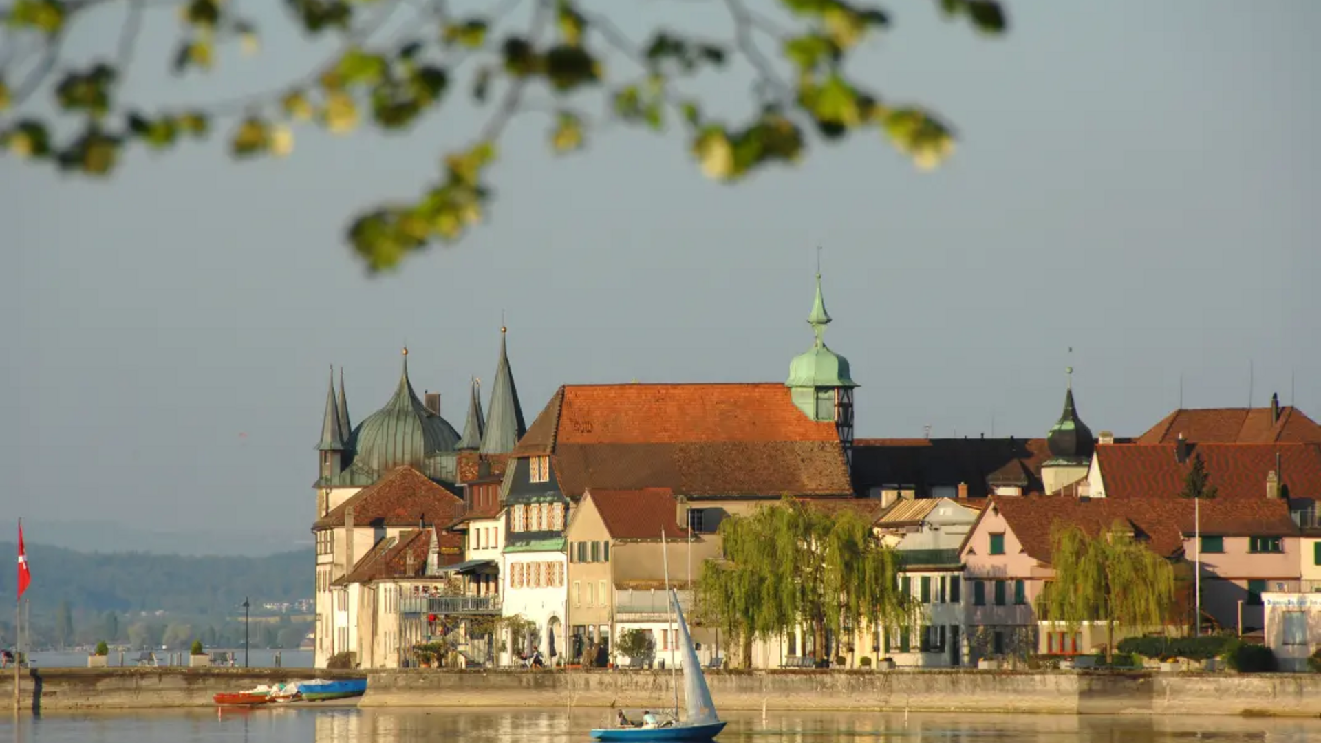 Historic lakeside buildings with sailboat and trees in the foreground