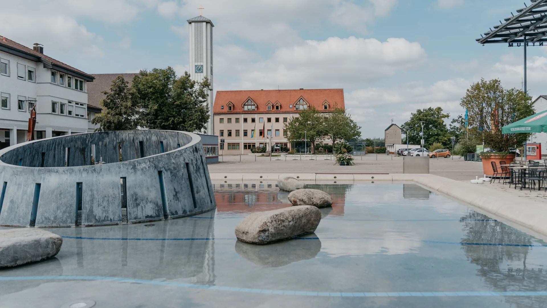 Moderner Brunnen mit Steinen und Kirchturm im Hintergrund bei bewölktem Himmel