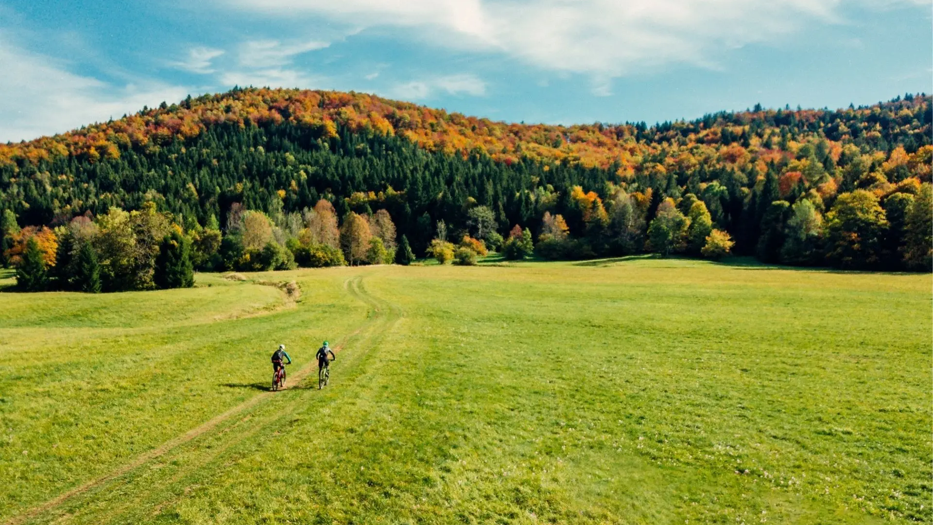 Two cyclists riding on a trail through a green meadow with autumn-colored hills