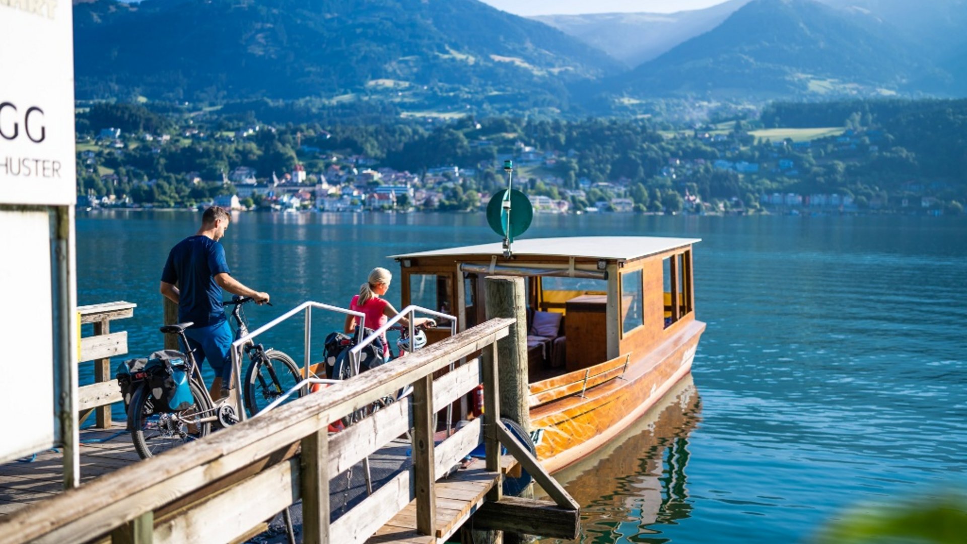 Two people with bicycles boarding a boat on a lake with mountain view