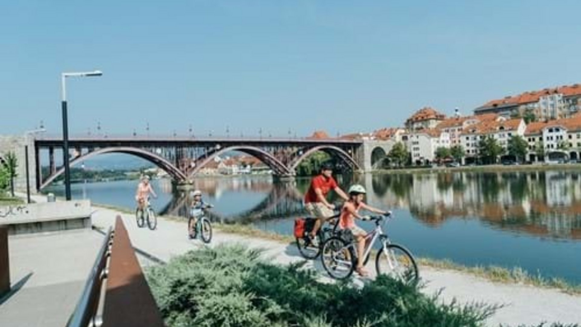 Family cycling along river with bridge and houses in background