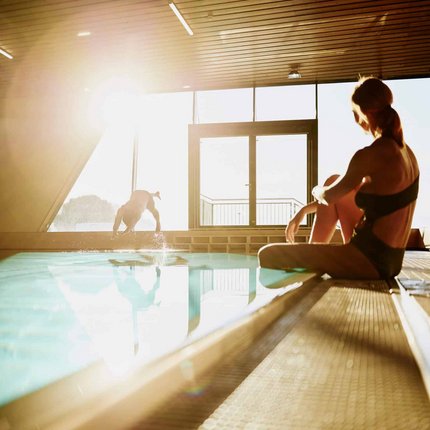 Woman sitting poolside indoors while person dives into the water