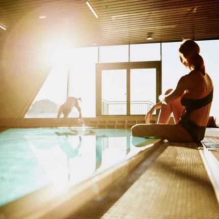 Woman sitting poolside indoors while person dives into the water