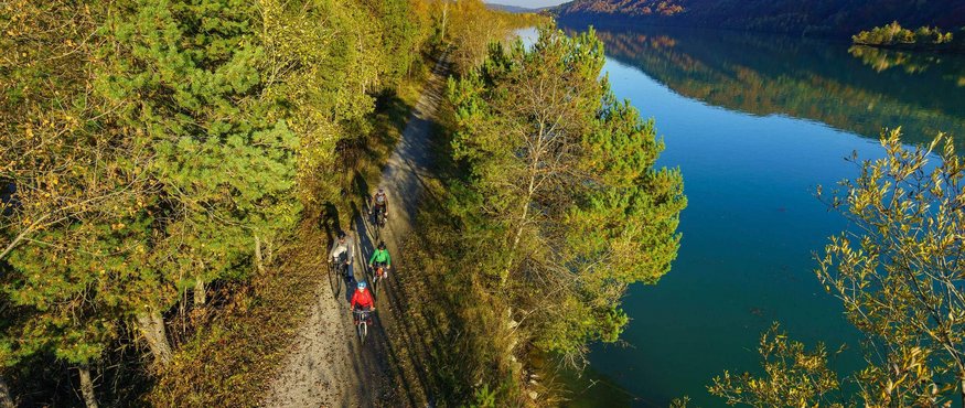 Cyclists on forest path next to calm river in autumn landscape