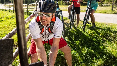 Mann holt Wasser mit Händen, zwei Radfahrer schauen zu, Berglandschaft im Hintergrund