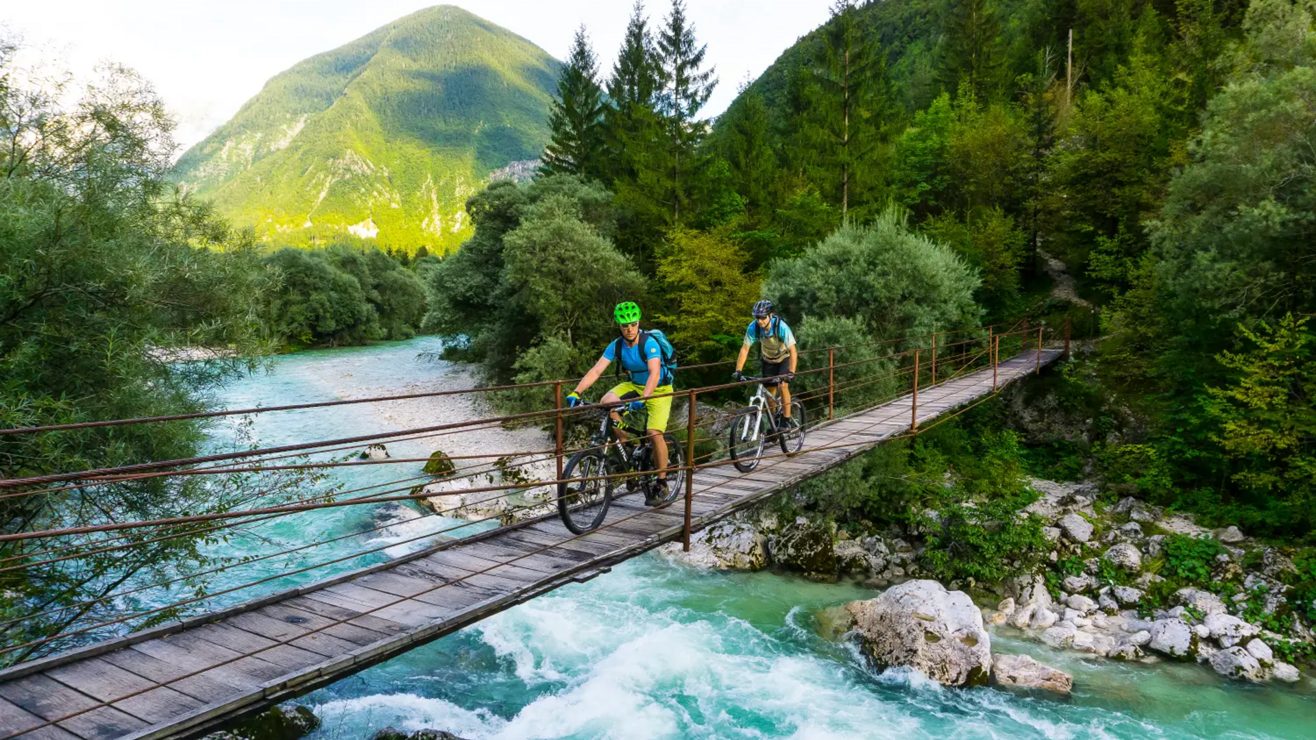 Two cyclists cross a suspension bridge over a rushing river in the mountains