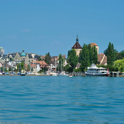 View of a lakeside promenade with boats and historic buildings under a clear blue sky