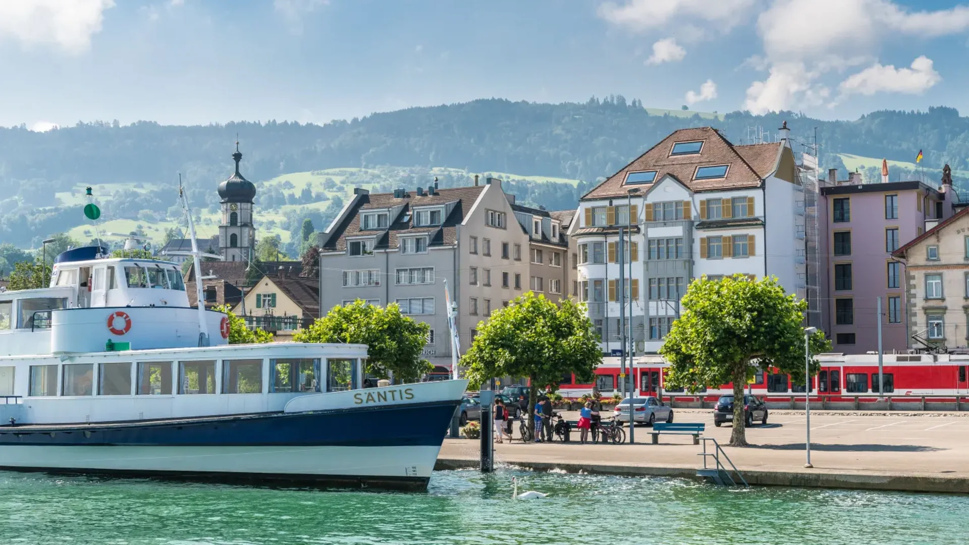 Boat docked by a lake shore with buildings and hills in the background