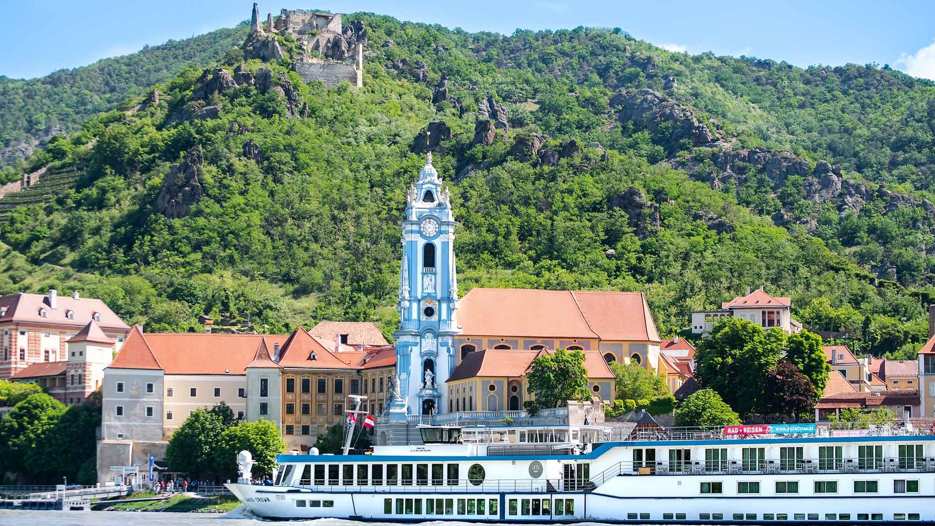 River cruise ship in front of town with church tower and forested hill