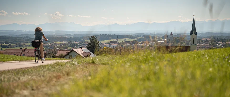 3 Gründe fürs Herbst-Radln in Inn-Salzach © Tourismusverein Inn-Salzach Fahrradfahrerin auf Landstraße mit Aussicht auf Dorf und Bergkulisse