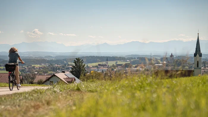 Fahrradfahrerin auf Landstraße mit Aussicht auf Dorf und Bergkulisse