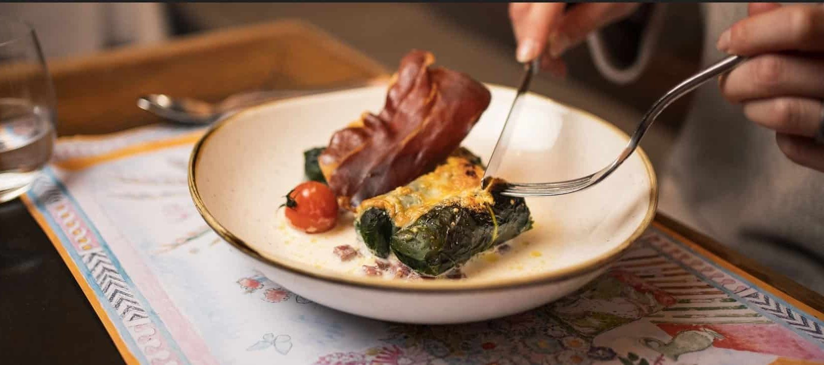 Person eating stuffed chard rolls with ham and tomato using fork and knife