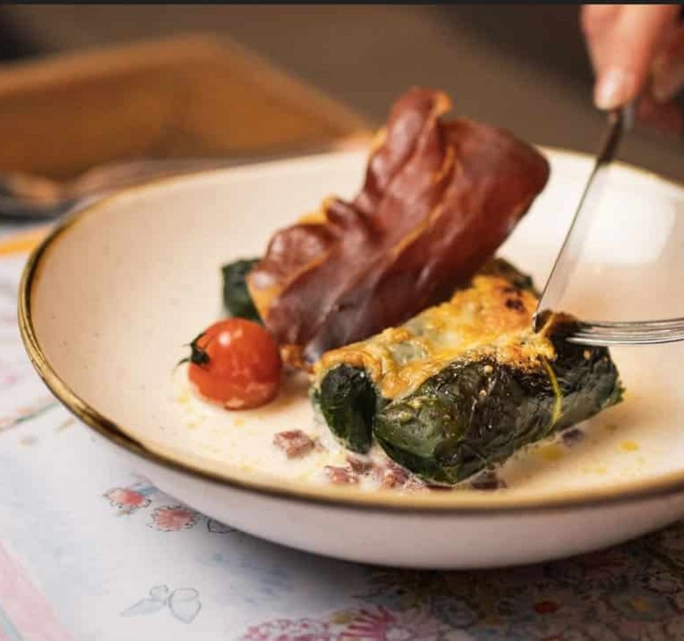 Person eating stuffed chard rolls with ham and tomato using fork and knife