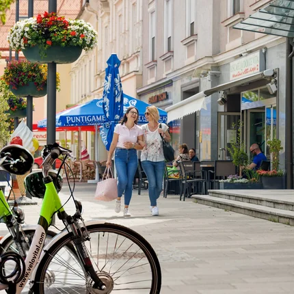 Two women laughing and walking on a lively street with bikes and flowers