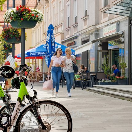 Two women laughing and walking on a lively street with bikes and flowers